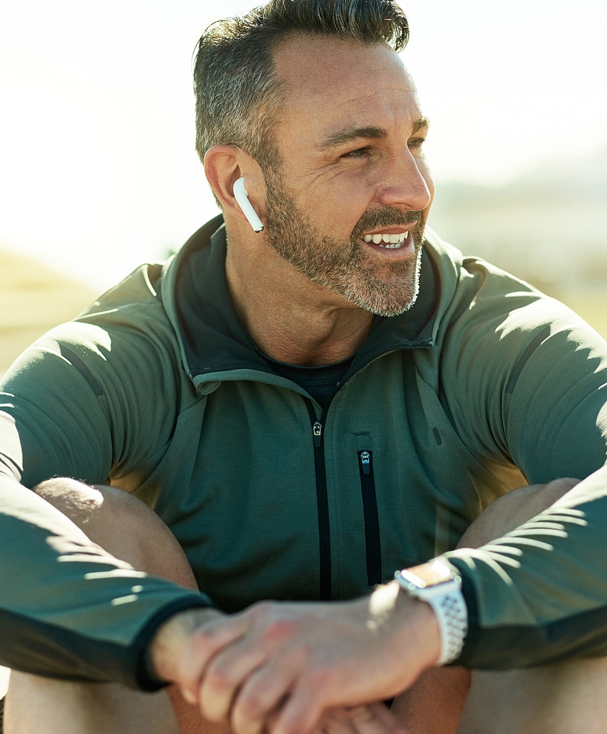 Smiling man wearing earbuds outdoors, enjoying sunlight.