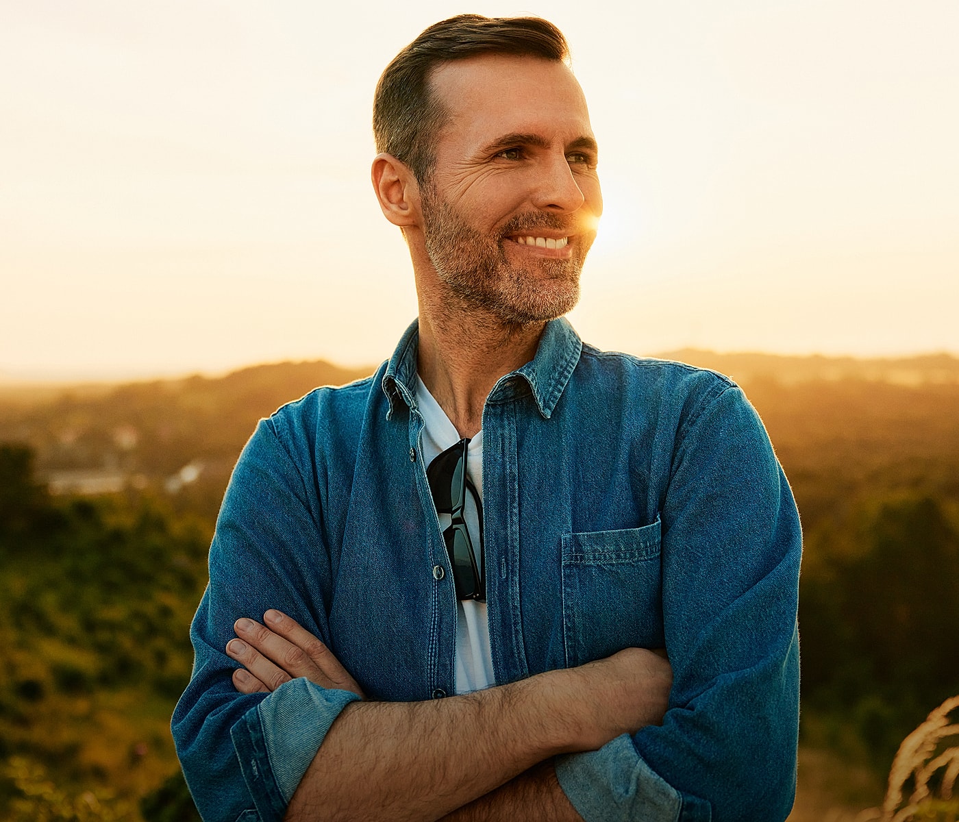 Man smiling in denim shirt at sunset.